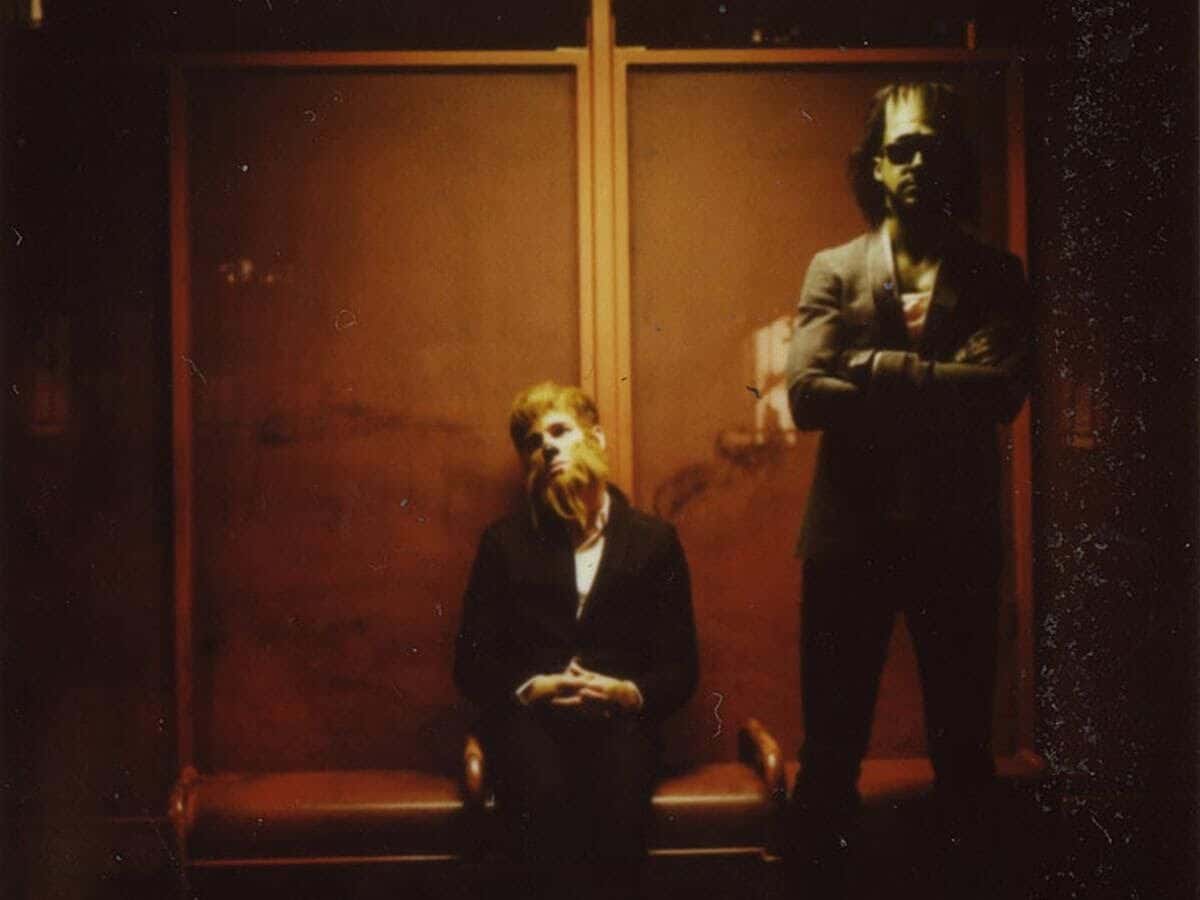 Foto de prensa de Dead Man's Bones: Ryan Gosling disfrazado de Frankenstein, de pie, y Zach Shields con máscara de hombre lobo, sentado, en un pasillo con paredes de madera y luz cálida