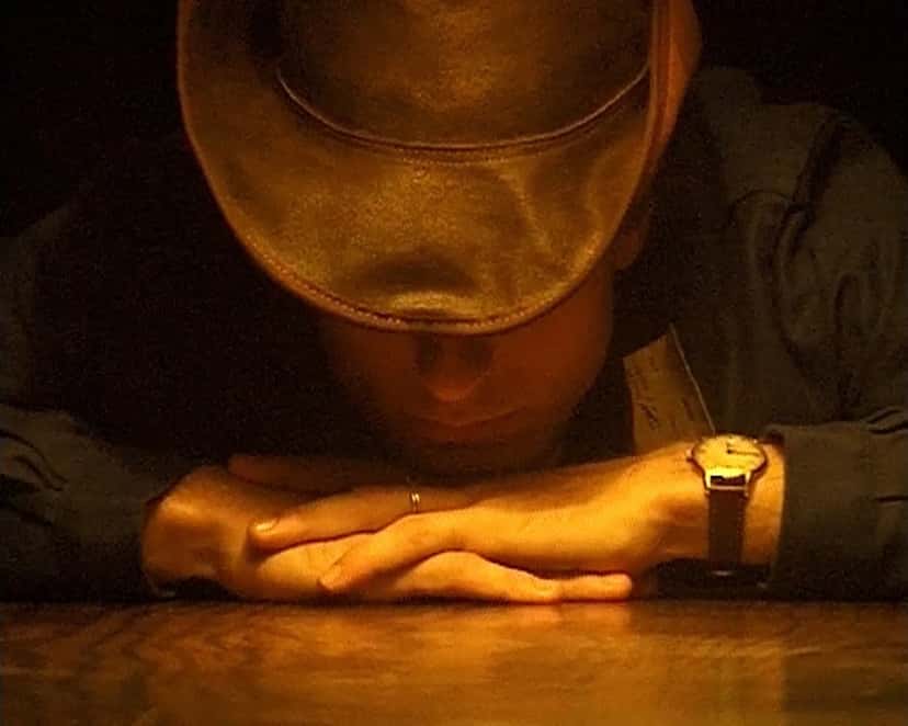 Escena del video oficial de «Forgetful Angel» de DJ Python: un hombre con sombrero de cowboy apoya la cabeza sobre sus manos cruzadas en una mesa, con luz cálida