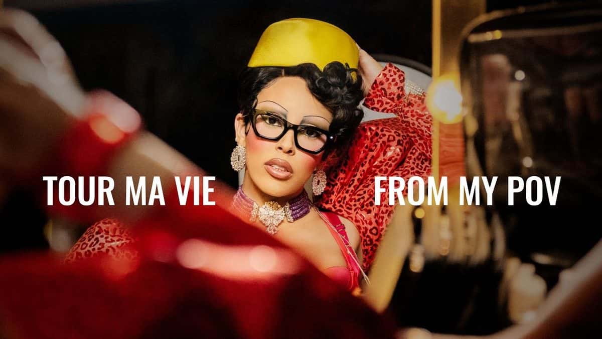 Doja Cat, con anteojos negros, gorro amarillo y saco rojo, frente a un espejo de camarín, en la imagen oficial de su documental con el texto «Tour Ma Vie from my POV»