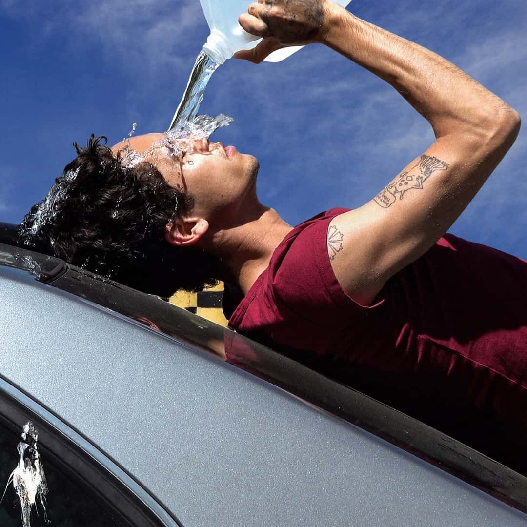 Portada de «How To Quit Smoking», de Dominic Fike: el músico, con remera bordó, se tira agua de un bidón sobre la cara, acostado sobre el techo de un auto gris bajo un cielo azul