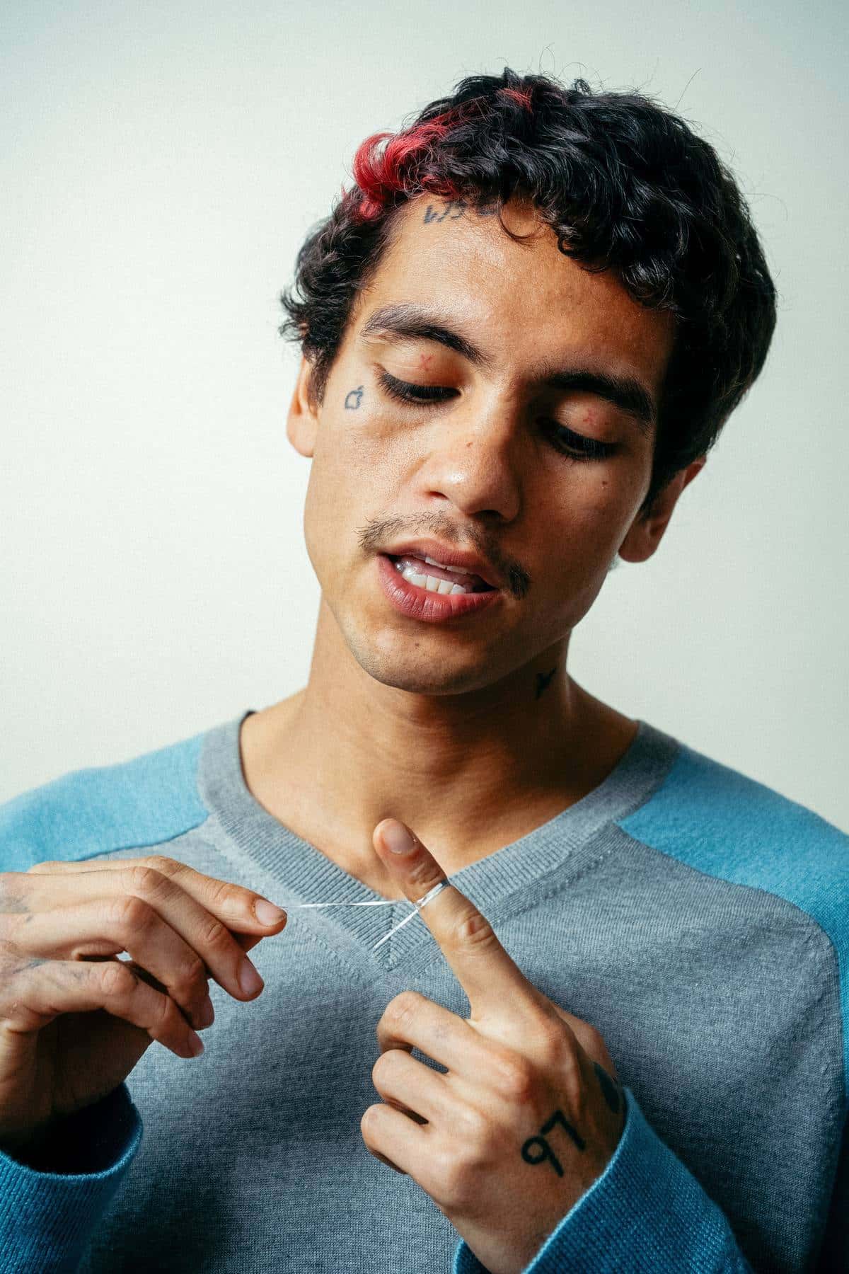 Dominic Fike, con pelo rulos oscuros con mechones rojos y un buzo gris y celeste, sostiene un hilo entre los dedos frente a un fondo claro, en una foto de prensa oficial
