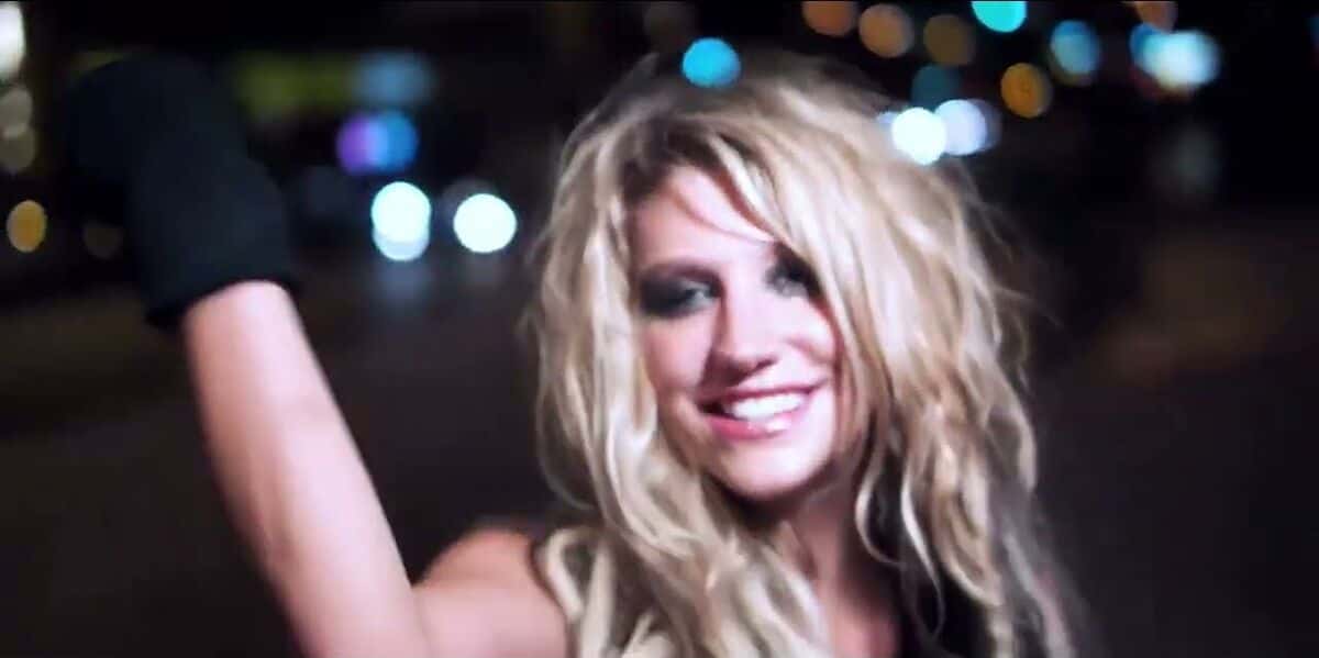 Fotograma del video oficial de «Backstabber»: Kesha, rubia y despeinada, sonríe con un guante negro y el brazo en alto en una calle de noche