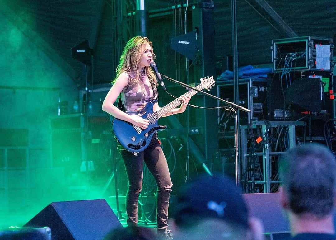 The Warning en vivo: una de las hermanas Villarreal toca una guitarra azul y canta frente al micrófono, con luces verdes y humo sobre el escenario, en junio de 2022 (foto de archivo). Foto: Tabercil, CC BY-SA 3.0, vía Wikimedia Commons