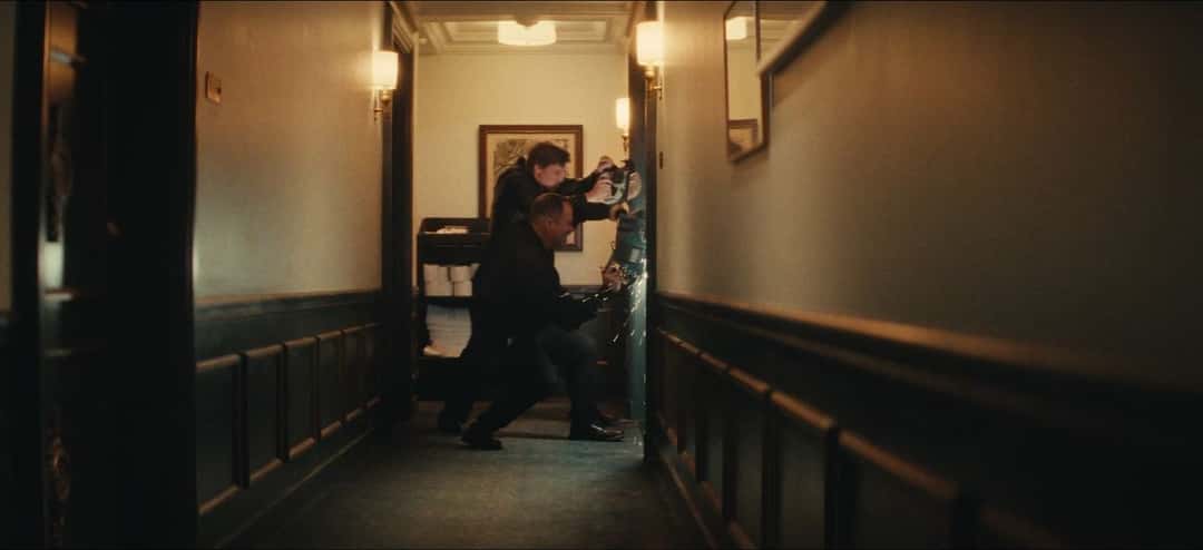 Fotograma del teaser de «Tarantino»: en un pasillo de hotel con paredes de madera y apliques encendidos, dos figuras de negro forcejean junto a una puerta