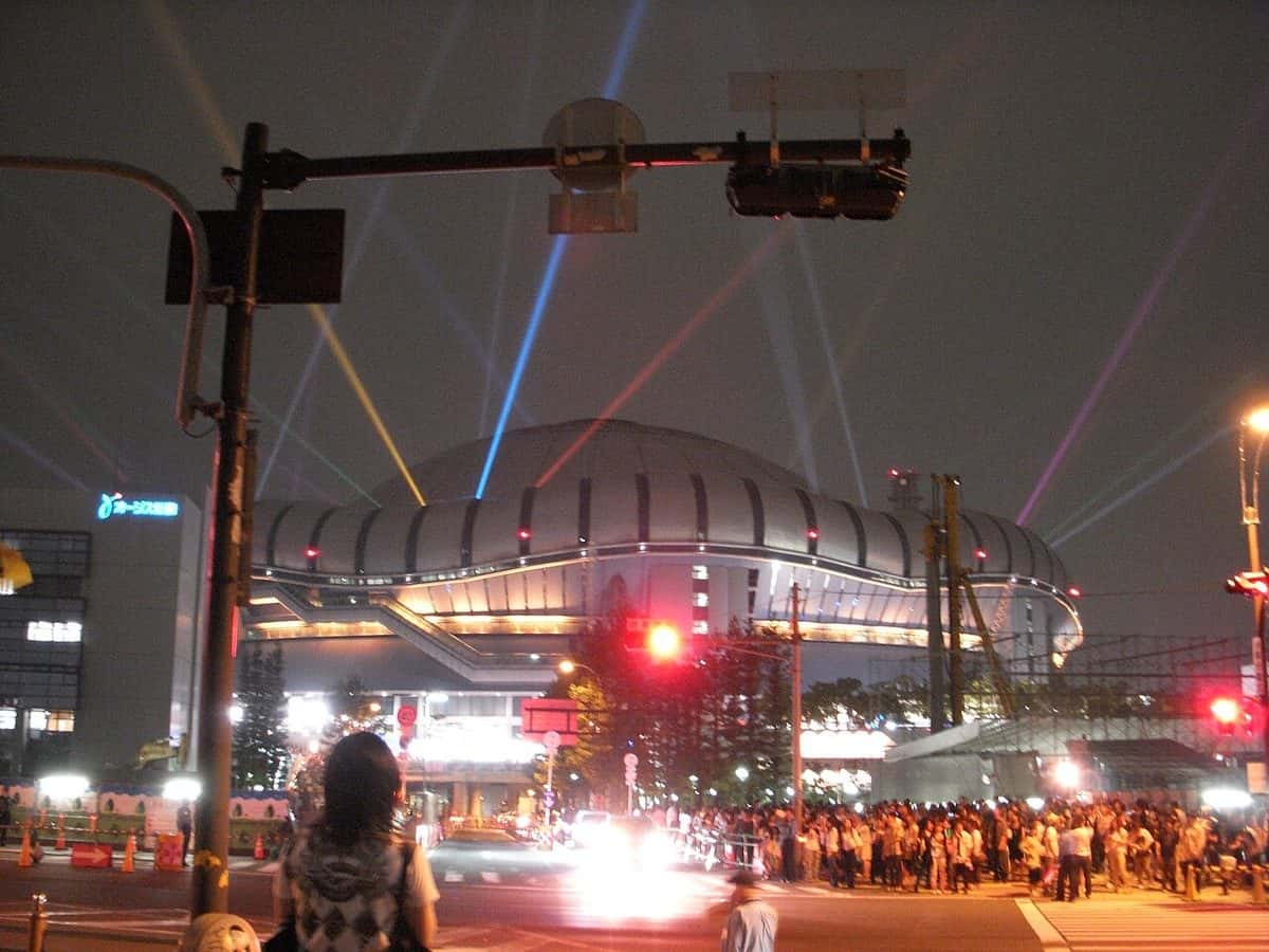 El Kyocera Dome de Osaka de noche, con rayos láser de colores que salen hacia el cielo y una multitud esperando en la vereda de enfrente (imagen del lugar de la ceremonia)