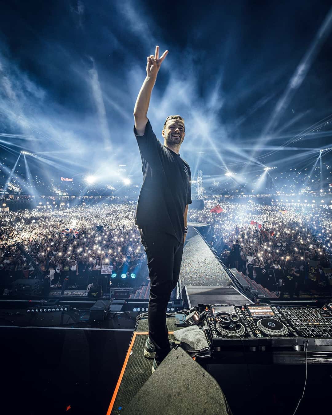 Martin Garrix de espaldas al público en un festival, con la mano en alto haciendo el signo de la paz, sobre la cabina de DJ, frente a una multitud iluminada por luces azules (foto de archivo)