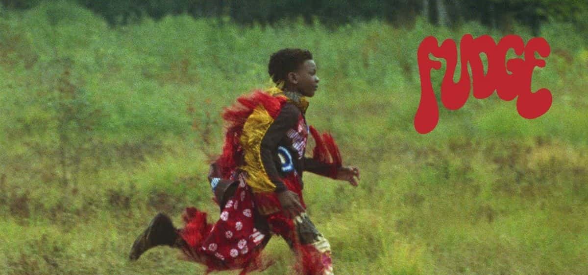 Fotograma del video oficial de «Summer Clothes»: una persona con un traje de flecos rojos, amarillos y blancos corre por un pastizal verde, con la palabra «FUDGE» en letras rojas arriba a la derecha