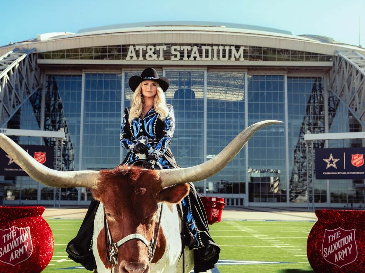 Miranda Lambert, con sombrero negro y camisa bordada en azul, montada sobre un toro de cuernos largos frente al AT&T Stadium, entre calderos rojos del Ejército de Salvación