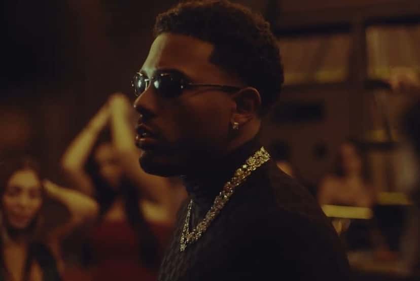 Myke Towers en el video oficial de «Te Imagino»: de perfil, con anteojos de sol y una cadena dorada, en un bar con gente bailando detrás (fotograma del video oficial)