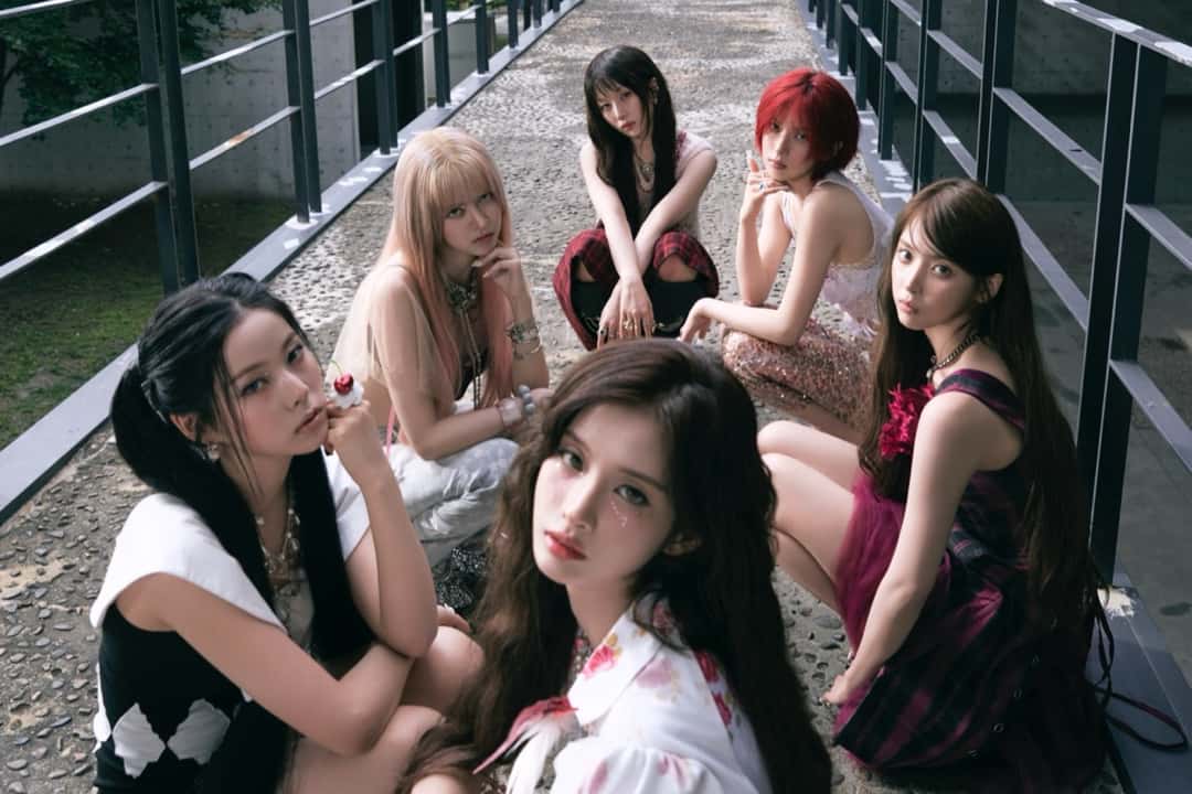 Las seis integrantes de NMIXX sentadas y agachadas sobre una pasarela de piedra con barandas de metal, en una foto conceptual oficial de «Strange Muse» (versión Strange)