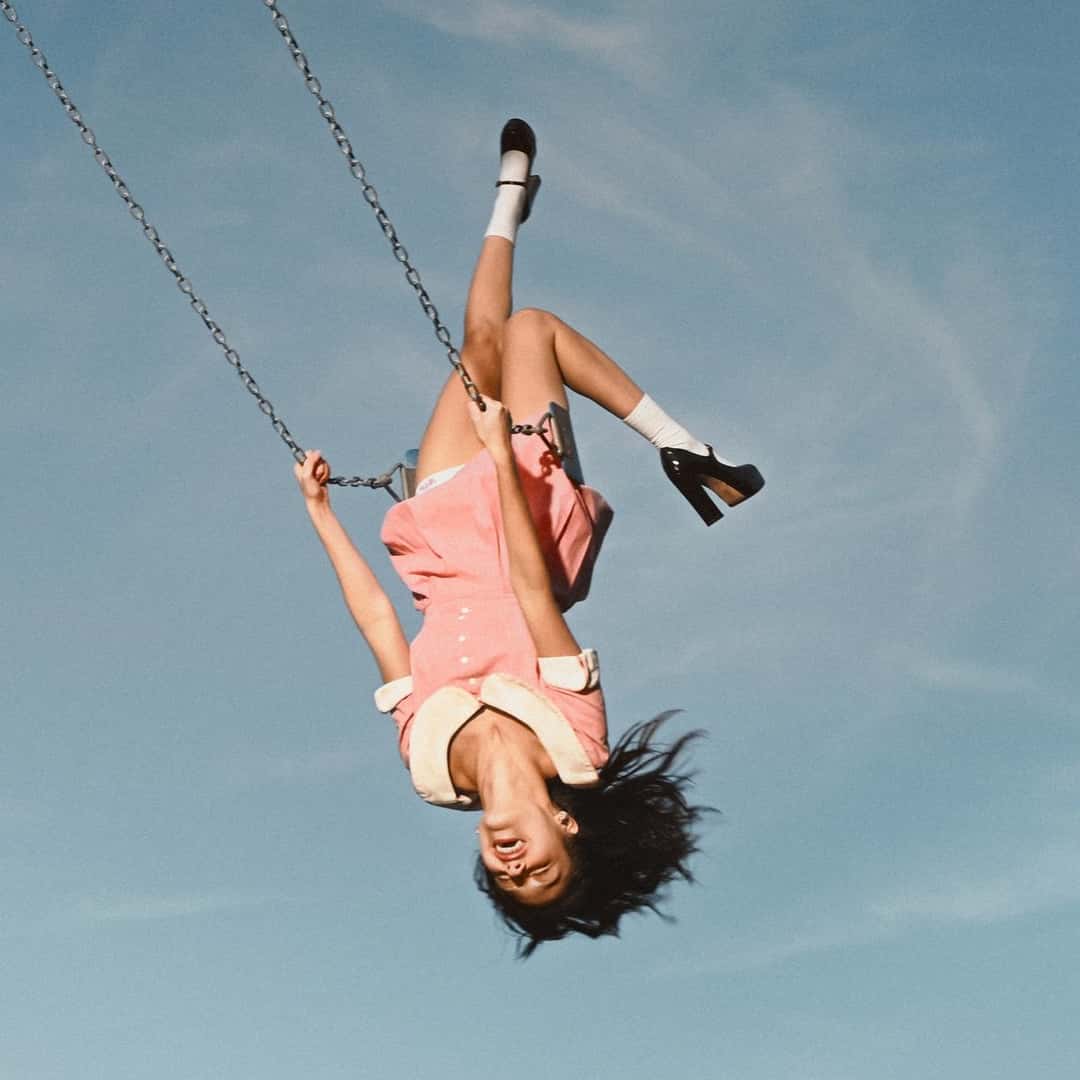 Portada de «you seem pretty sad for a girl so in love», de Olivia Rodrigo: la cantante, con vestido rosa y zapatos negros, cuelga cabeza abajo de una hamaca contra un cielo celeste