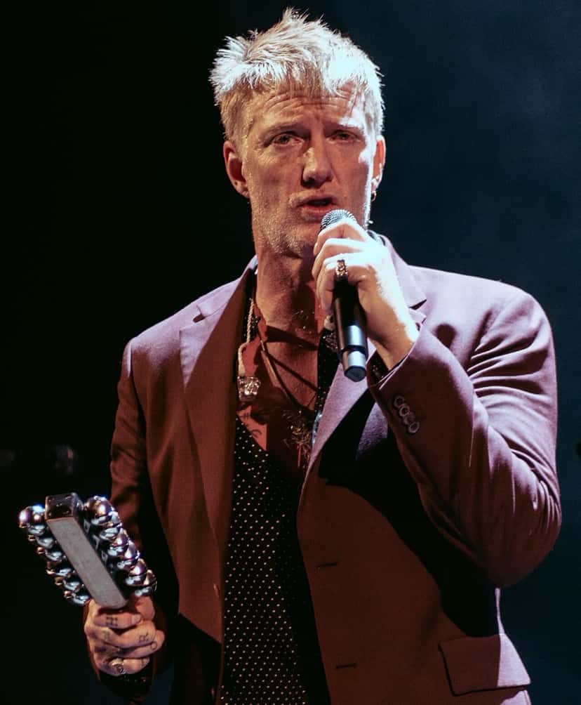 Josh Homme canta con un micrófono en la mano y una pandereta en la otra, con saco bordó y camisa oscura, durante un show (foto de archivo, octubre de 2025). Foto: Raph_PH, CC BY 4.0, vía Wikimedia Commons