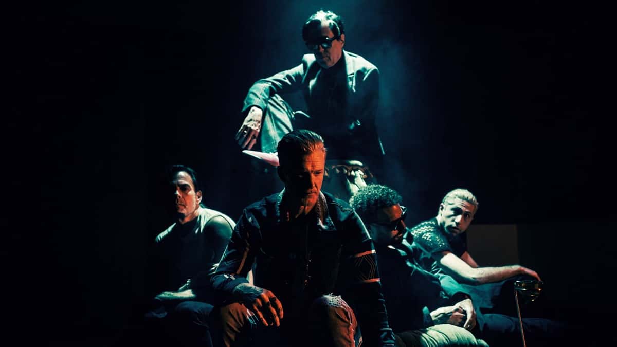 Foto de prensa de Queens of the Stone Age para «Perfecth»: los cinco integrantes en penumbra, uno sentado adelante y otro de anteojos oscuros arriba de un sillón, bajo una luz verde azulada