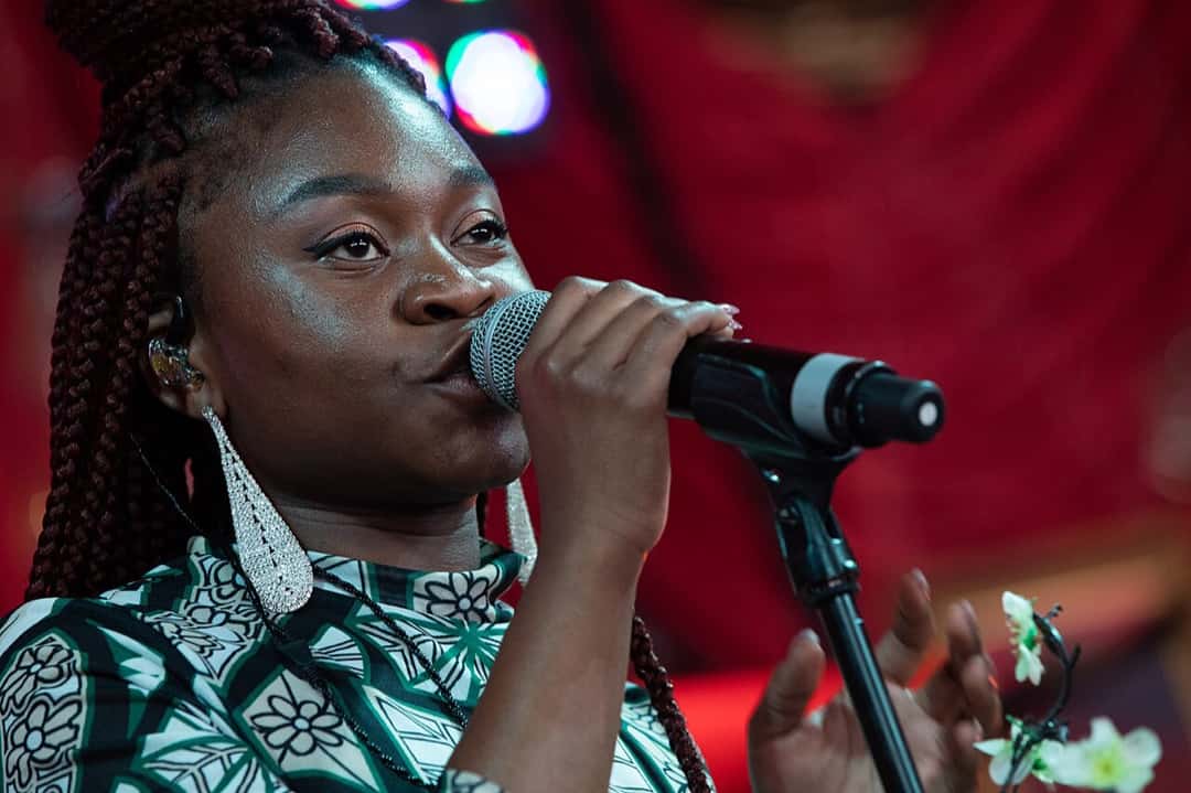 Sampa the Great canta con el micrófono en la mano, con trenzas bordó, aros largos plateados y una prenda con estampado verde y blanco, sobre un escenario con fondo rojo, en el festival de Glastonbury de 2019 (foto de archivo). Foto: Simoncromptonreid, CC BY-SA 4.0, vía Wikimedia Commons