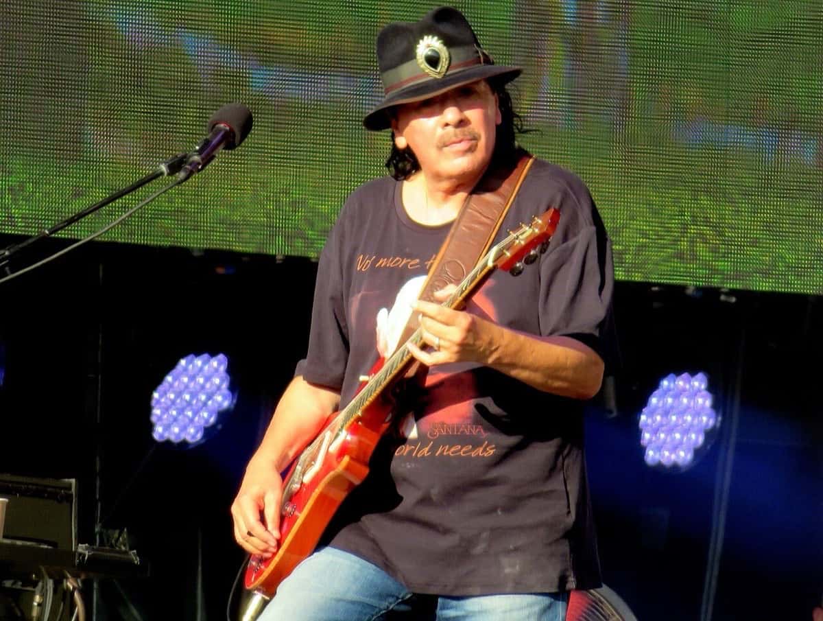 Carlos Santana toca una guitarra eléctrica roja con sombrero negro y remera oscura, frente a una pantalla verde, en un show al aire libre en Londres (foto de archivo, agosto de 2018)