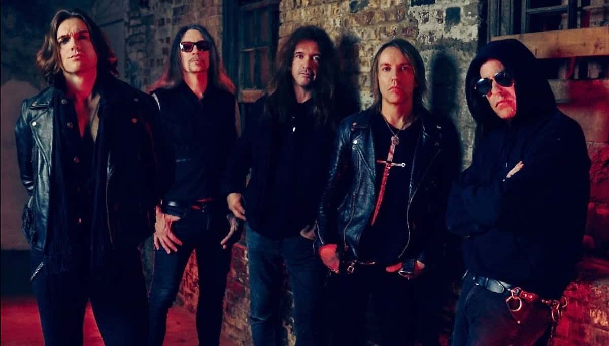 Foto de prensa de Skid Row con su nueva formación: cinco músicos de pelo largo y ropa negra, varios con campera de cuero y anteojos oscuros, posan contra una pared de ladrillos con luz roja