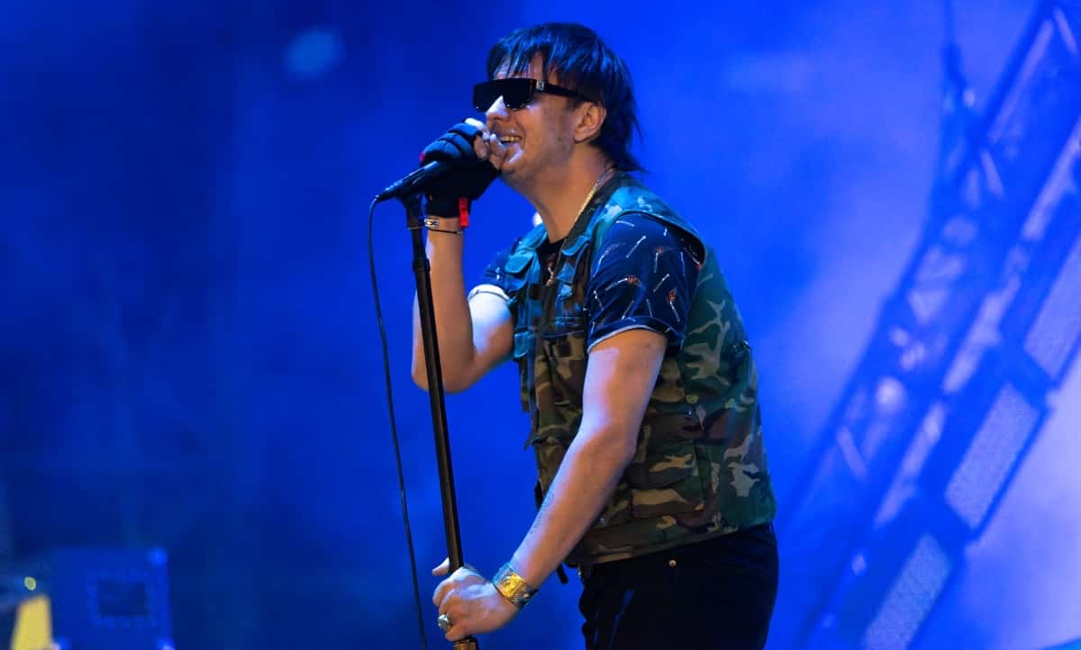 Julian Casablancas, cantante de The Strokes, en vivo en el festival NOS Alive de Lisboa en julio de 2022: canta con anteojos oscuros y camisa de camuflaje, agarrado al pie del micrófono, frente a luces azules (foto de archivo). Foto: Raph_PH, CC BY 2.0, vía Wikimedia Commons