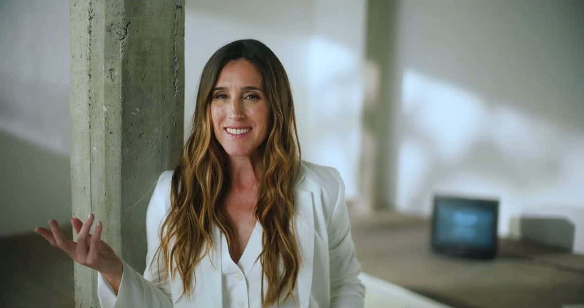 Fotograma del video oficial de «Vale la pena»: Soledad sonríe con saco blanco y pelo largo suelto, apoyada junto a una columna de hormigón en un ambiente luminoso