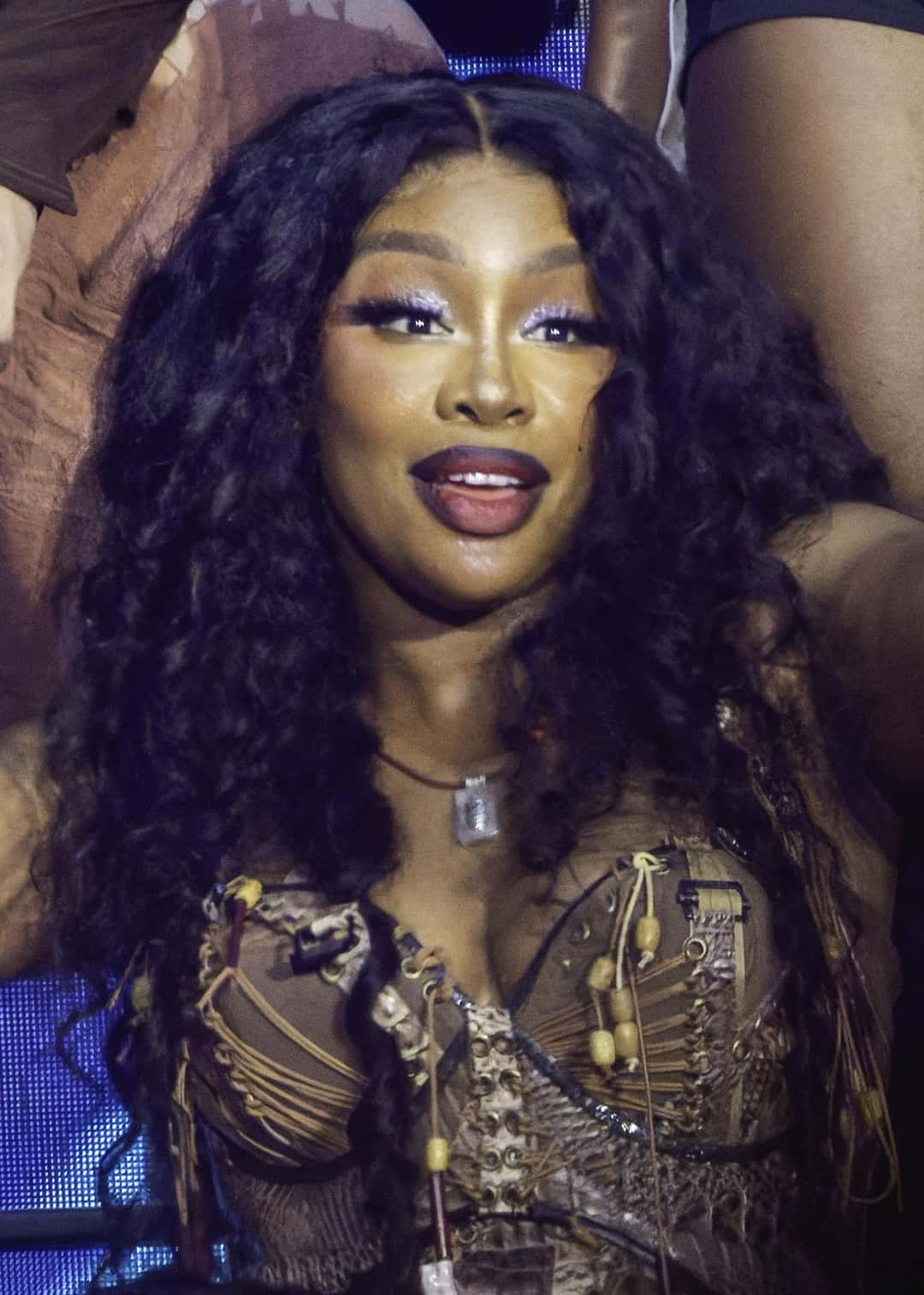 SZA sonríe en el escenario del festival de Glastonbury, con el pelo largo y rizado y un corsé marrón con tiras y cuentas de madera (foto de archivo, junio de 2024). Foto: Raph_PH, CC BY 2.0, vía Wikimedia Commons