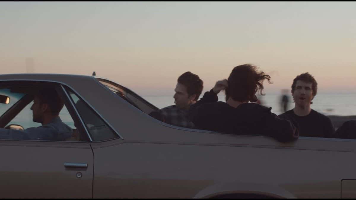 Los integrantes de The Neighbourhood, a contraluz, viajan sentados arriba del respaldo de un auto descapotable color beige frente al mar al atardecer, en una imagen del video oficial de «Lovebomb»