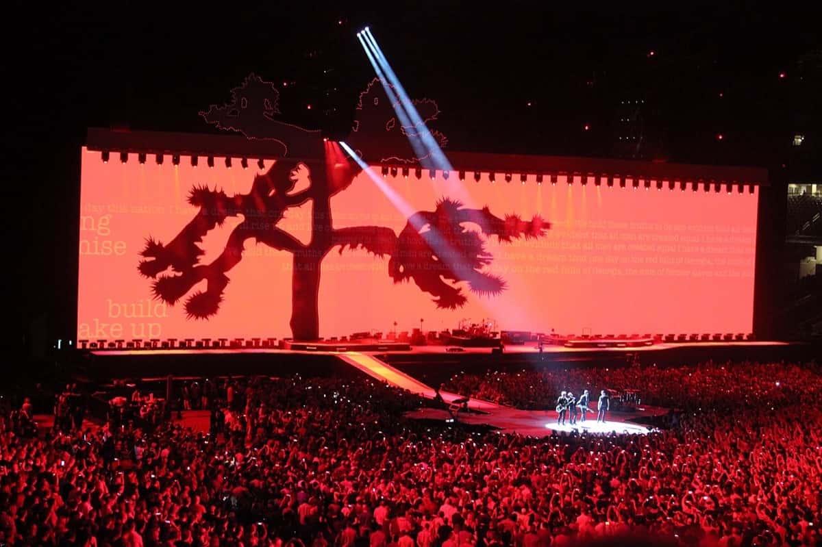 U2 toca en una pasarela en medio del público de un estadio, con una enorme pantalla roja de fondo que muestra la silueta de un árbol de Josué, en mayo de 2017 (foto de archivo)