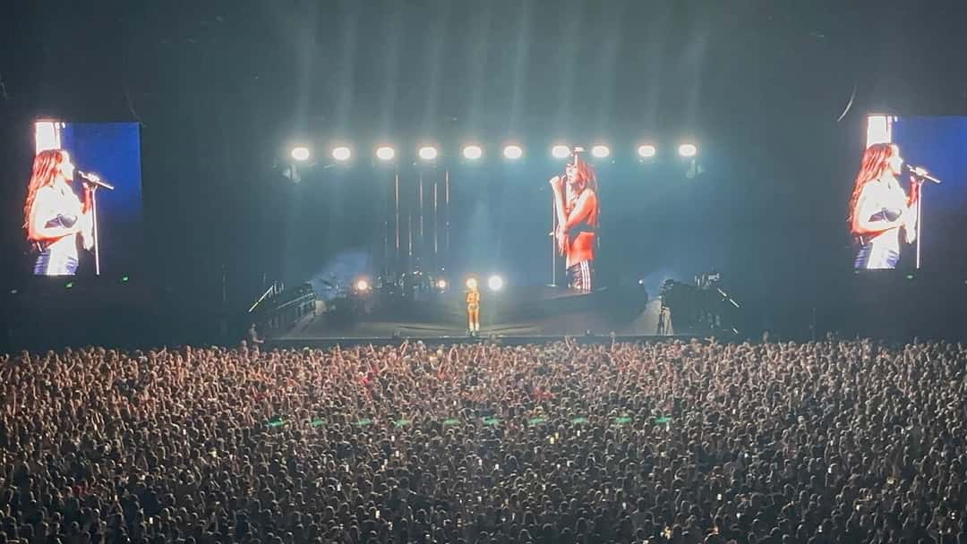 Foto de archivo de Tate McRae en el escenario durante un show en Adelaida, Australia, en noviembre de 2024: la cantante aparece en las pantallas gigantes a los costados mientras el estadio lleno la mira bajo una fila de reflectores blancos. Foto: Avatar5991, Wikimedia Commons, CC BY-SA 4.0