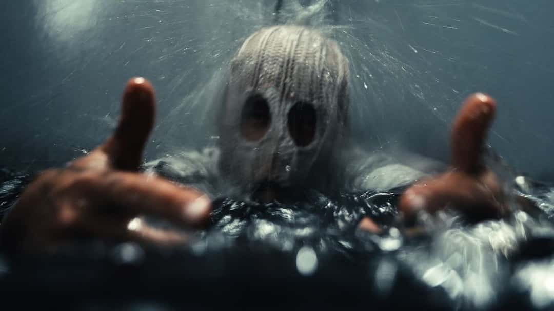 Fotograma de archivo del video oficial de «EARNËD IT»: alguien con una máscara tejida de color crema sale del agua oscura con los brazos estirados hacia la cámara