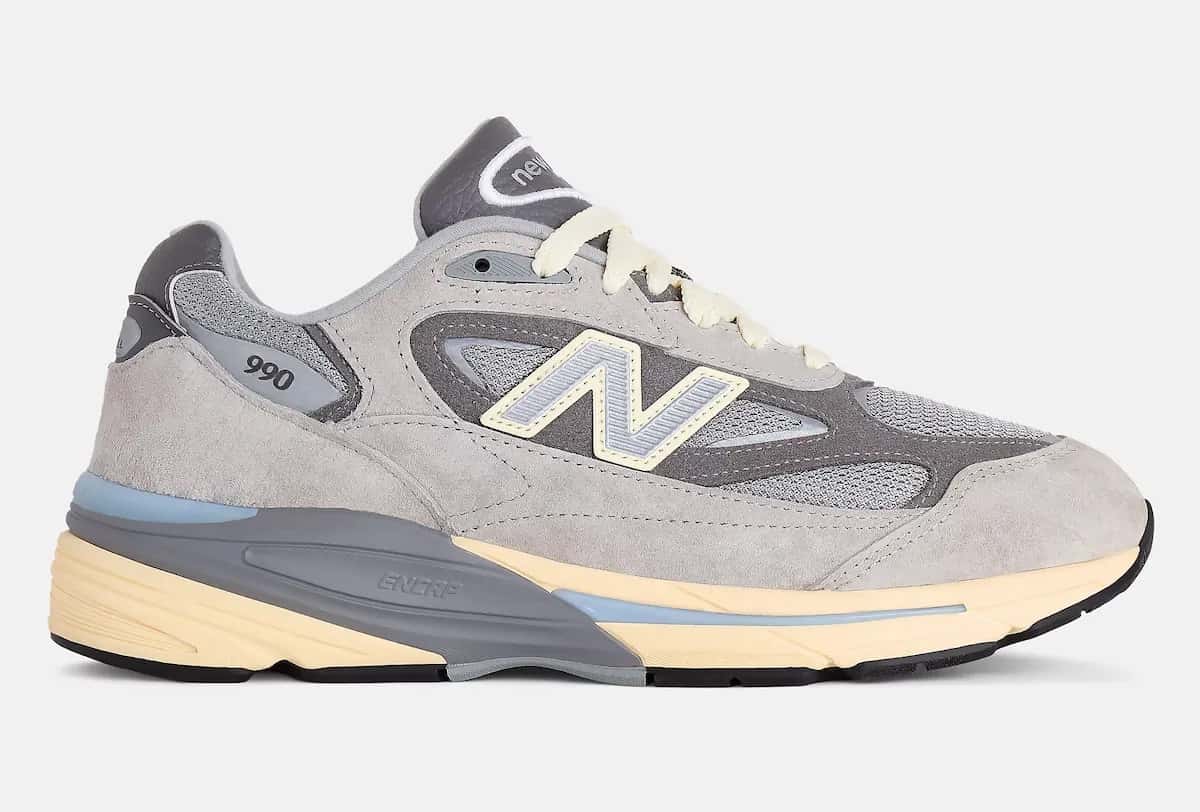 New Balance 990v7 Made in USA Kool Grey de perfil sobre fondo gris claro, con gamuza gris clara y oscura, malla gris, la N con relleno celeste y borde amarillo y la entresuela en gris y amarillo pálido