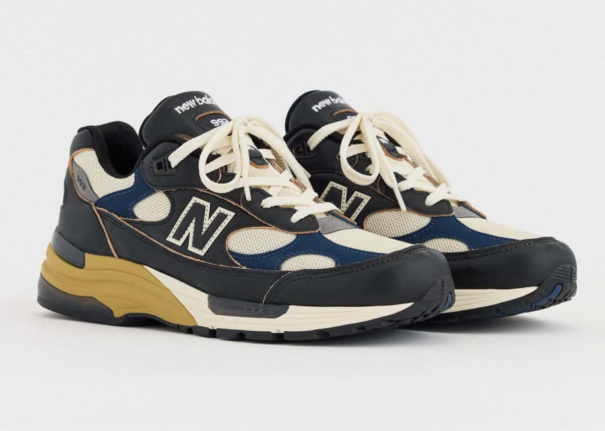 Par de New Balance 992 Made in USA Black/Afterglow en vista tres cuartos sobre fondo gris claro, con cuero negro de bordes tizados, malla crema, apliques azul marino, cordones crema y talón de la mediasuela en tono mostaza