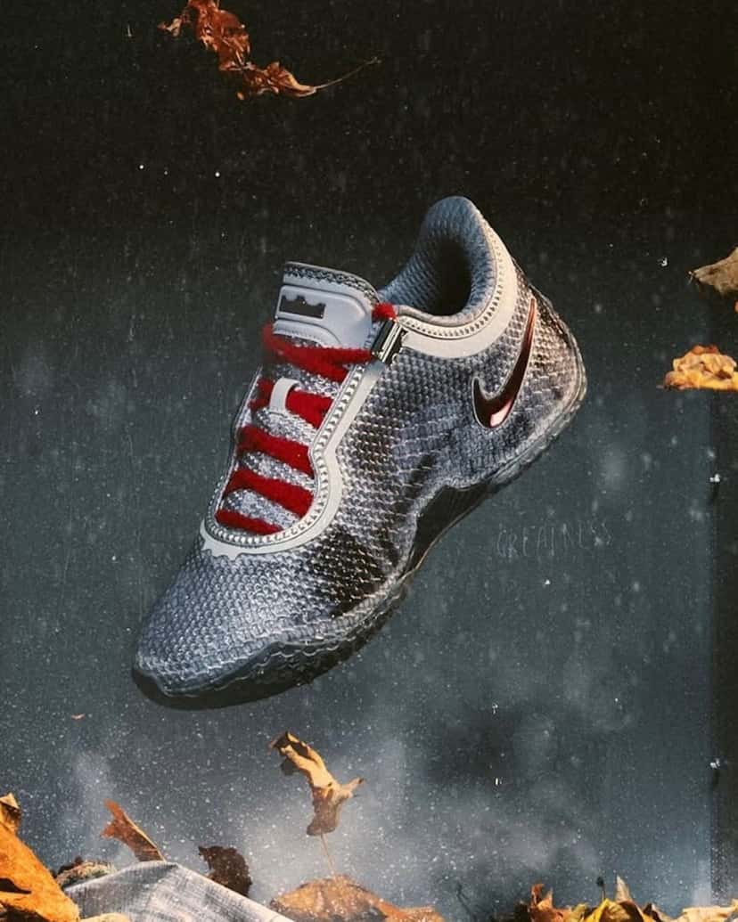 Nike LeBron 24 Storm Drain suspendido en el aire en una foto de campaña sobre un fondo oscuro con gotas y hojas secas, con upper tejido gris plateado con manchas oscuras, ribetes con tachas en el borde, cordones rojos de textura afelpada, swoosh rojo metalizado y el logo de la corona en la lengüeta