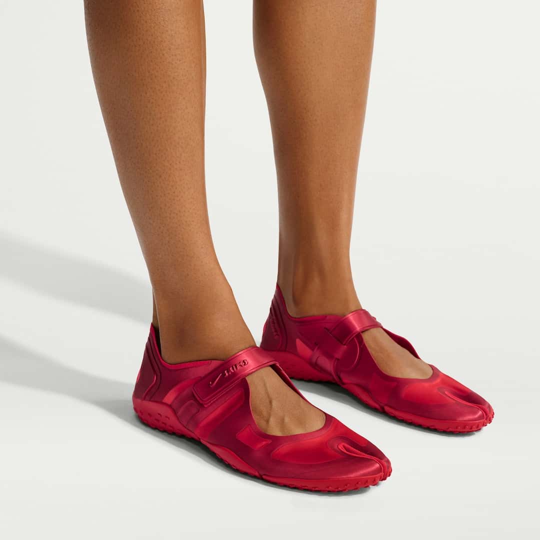 Pies con la NikeSKIMS Rift Satin University Red puesta sobre fondo blanco, con la punta dividida, la tira de abrojo sobre el empeine y la suela roja