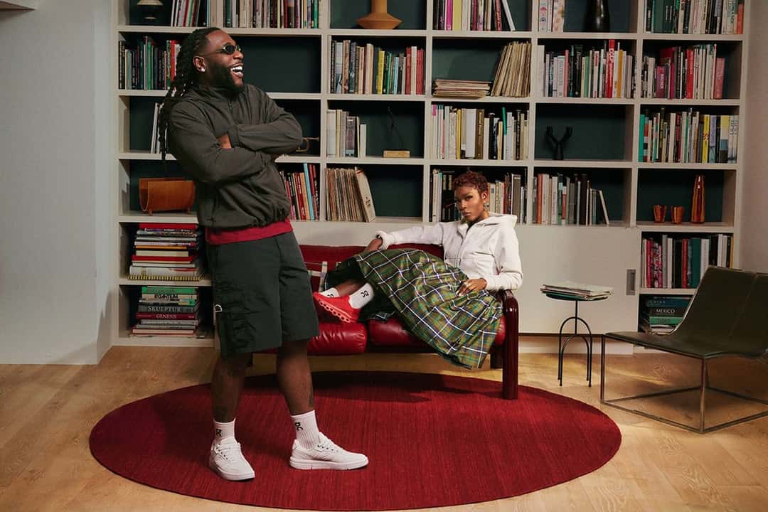 Burna Boy en una biblioteca con una alfombra roja, con campera oscura, bermuda y la On THE ROGER Clubhouse BB blanca, junto a una mujer sentada en un sillón con zapatillas rojas, en una foto de la campaña