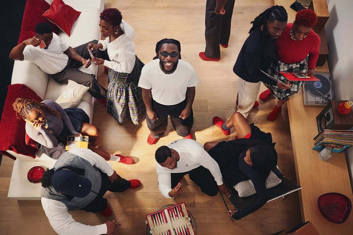 Burna Boy sentado en el centro de un grupo de once personas de su entorno, casi todos con zapatillas On Cloudmonster rojas, en una sala con piso de madera