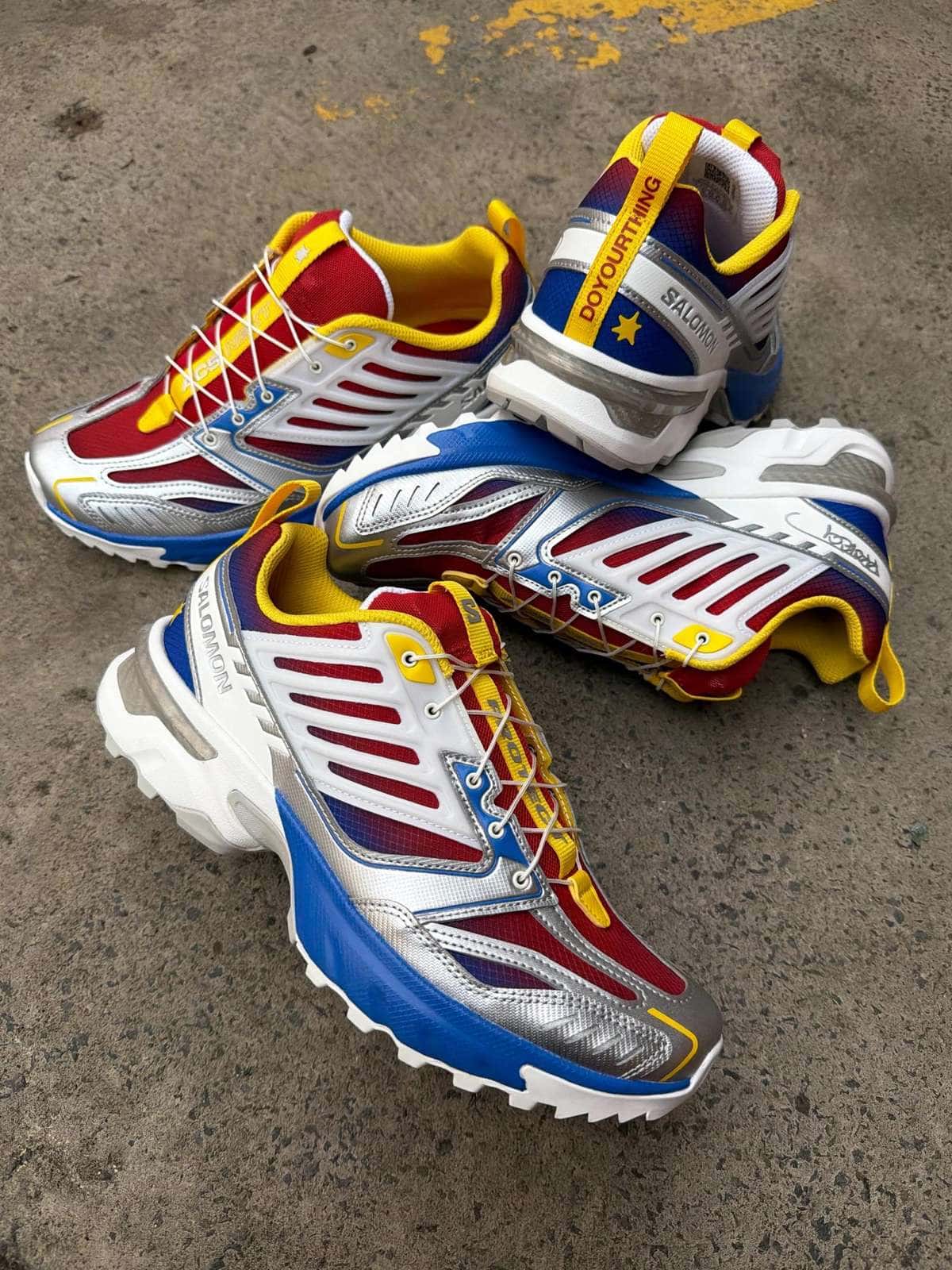 Par de Salomon ACS NEXT Slushie de PROTECT sobre el asfalto, con capellada roja y azul, refuerzos plateados, jaula blanca, detalles amarillos y suela azul y blanca