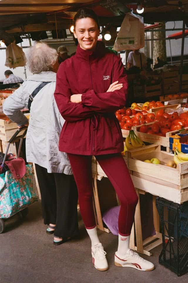 Una modelo con campera bordó y calzas de la colección Reebok x Rouje, con la Freestyle crema y medias blancas, en un puesto de frutas de París