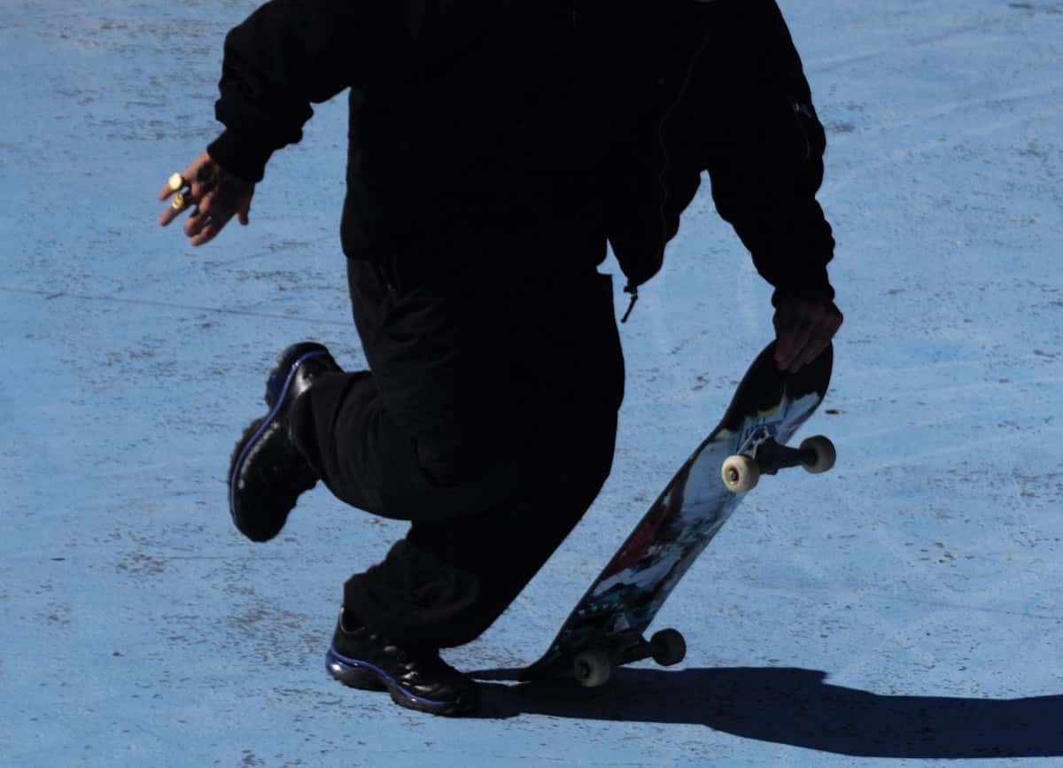 Foto de la muestra SYNC de Salomon: un skater vestido de negro pisa la cola de su tabla sobre el asfalto, con luz fuerte de sol