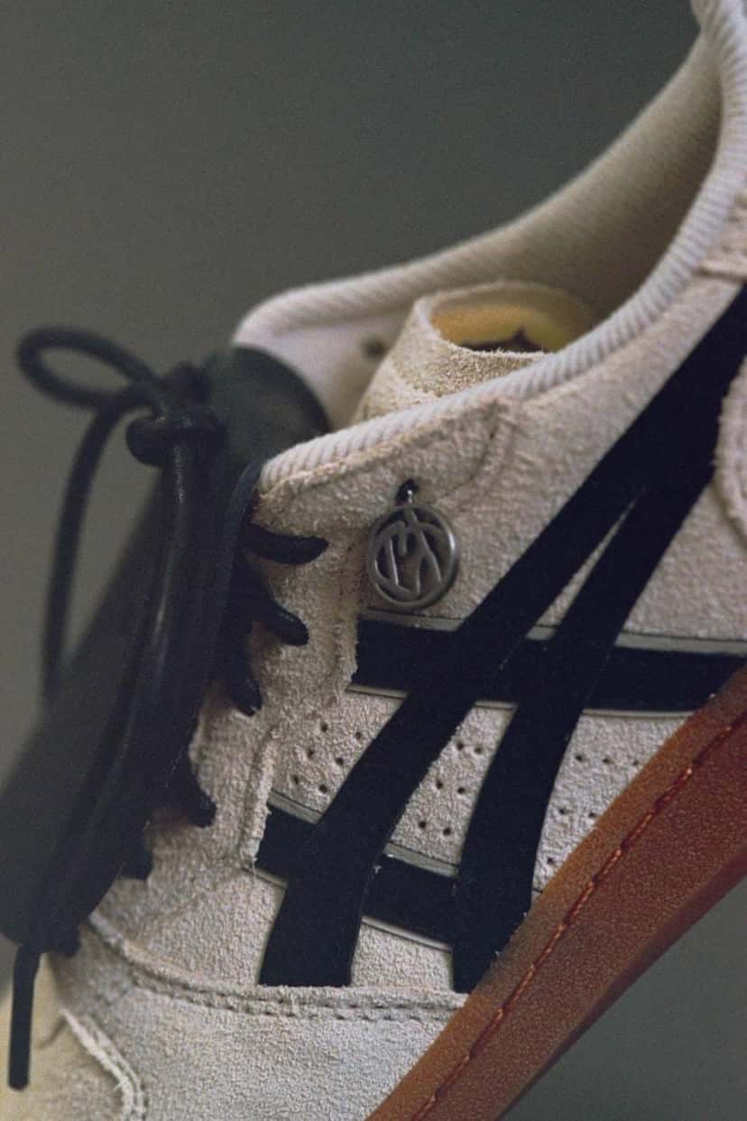 Lateral de la SOSHIOTSUKI x ASICS Skyhand OG con las rayas ASICS negras sobre la gamuza crema y el dije metálico colgando del ojal