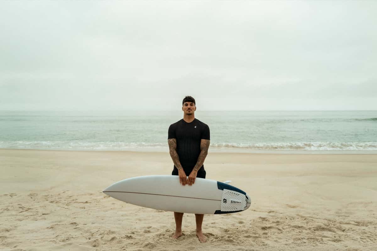Gabriel Medina, con la remera negra de la colección «Drop the Fear», sostiene una tabla blanca en una playa de arena con el mar de fondo