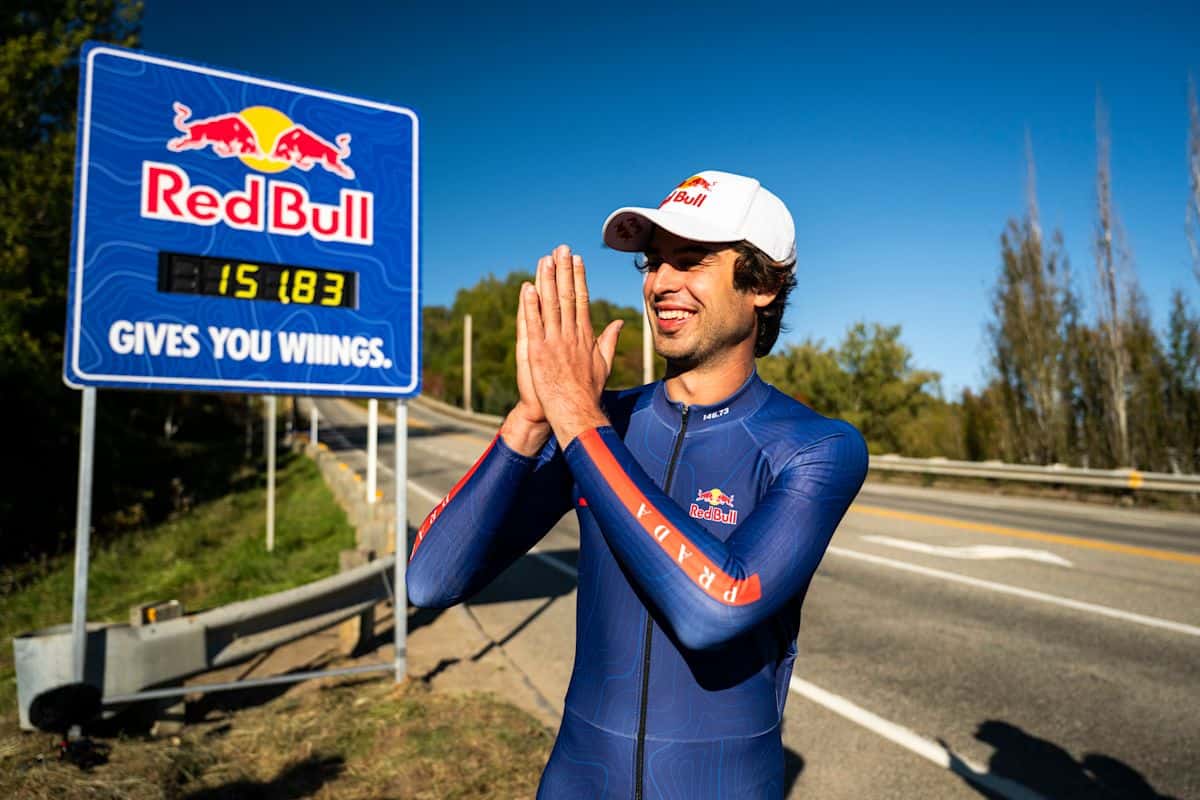 Diego Poncelet, con gorra blanca y traje aerodinámico azul, junta las manos y sonríe al costado de la ruta, junto a un cartel que marca 151.83