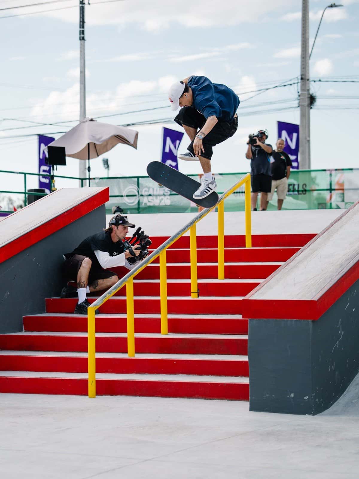 Angelo Caro, con buzo azul y gorra blanca, salta con la tabla en el aire junto a la baranda amarilla de la escalera roja de la pista de street de Luque durante la semifinal, con un camarógrafo agachado al costado